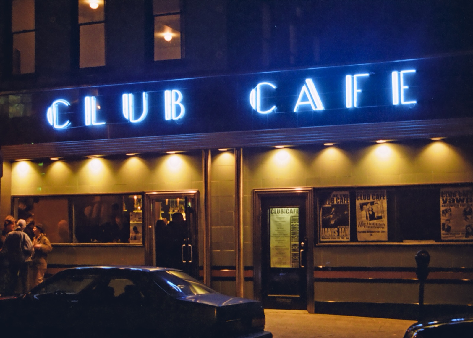 A blue neon Club Cafe sign glows over the venue's storefront at night, with show posters in the windows.